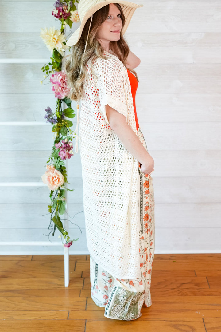Woman wearing a white crochet cover-up with a floral arrangement on a wooden floor.