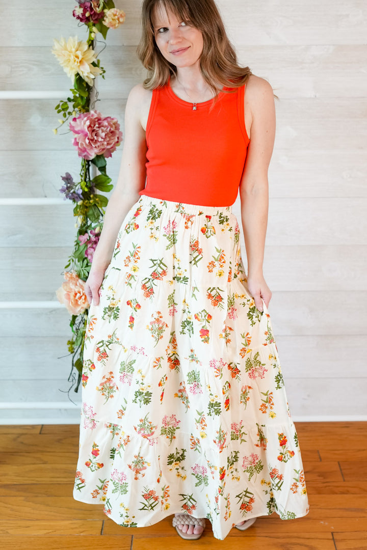 Woman wearing a bright red orange tank top top and vintage botanical floral skirt standing against a light wooden background with flowers.