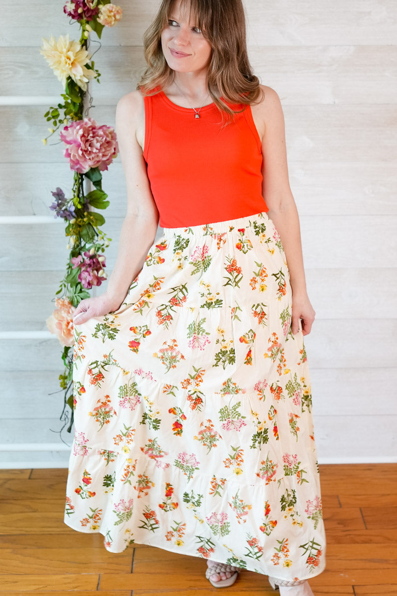 Woman wearing a red tank top and vintage floral maxi skirt standing against a white wall with decorative flowers.