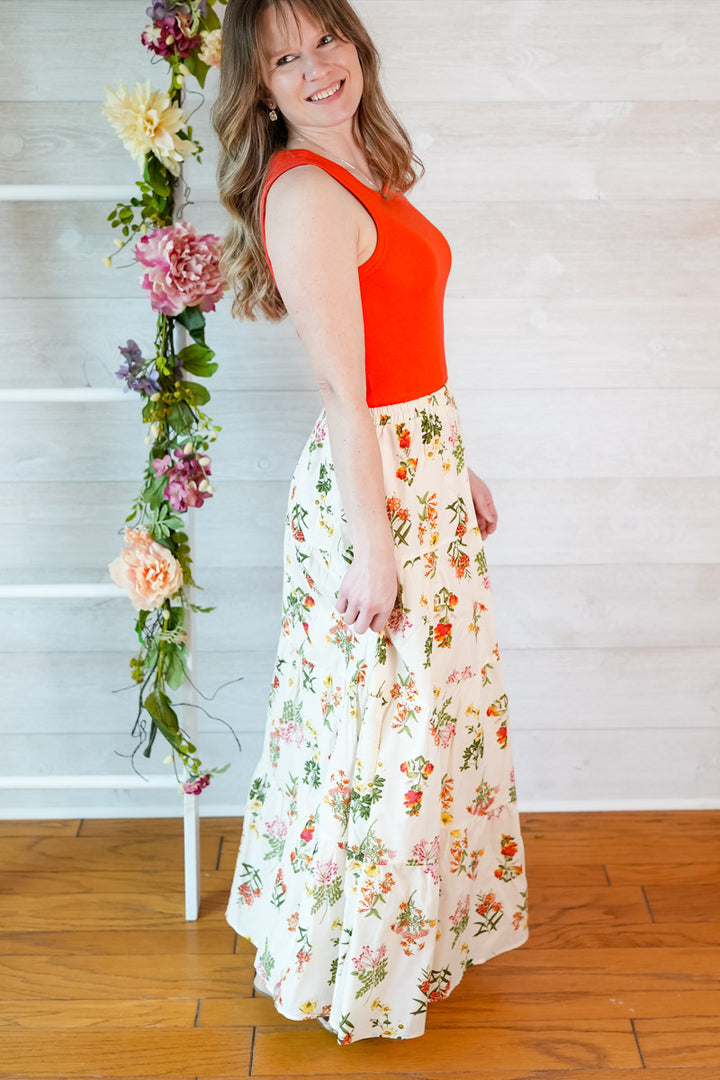Woman wearing an red orange tank top top and vintage floral skirt standing next to a floral arrangement indoors.