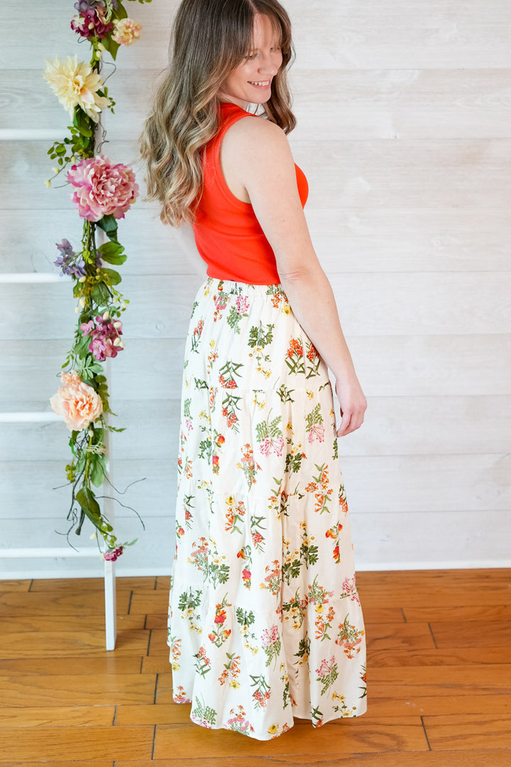 Woman wearing a red tank top top and vintage floral skirt standing next to a floral arrangement indoors.