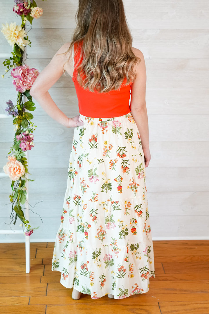 Woman wearing a red tank top and vintage floral skirt standing next to a floral arrangement against a white wall.