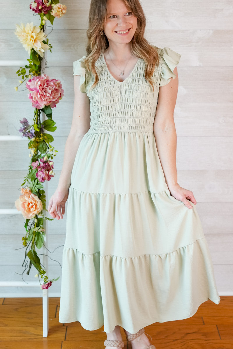 Woman wearing a light sage green dress standing next to a floral arrangement indoors.