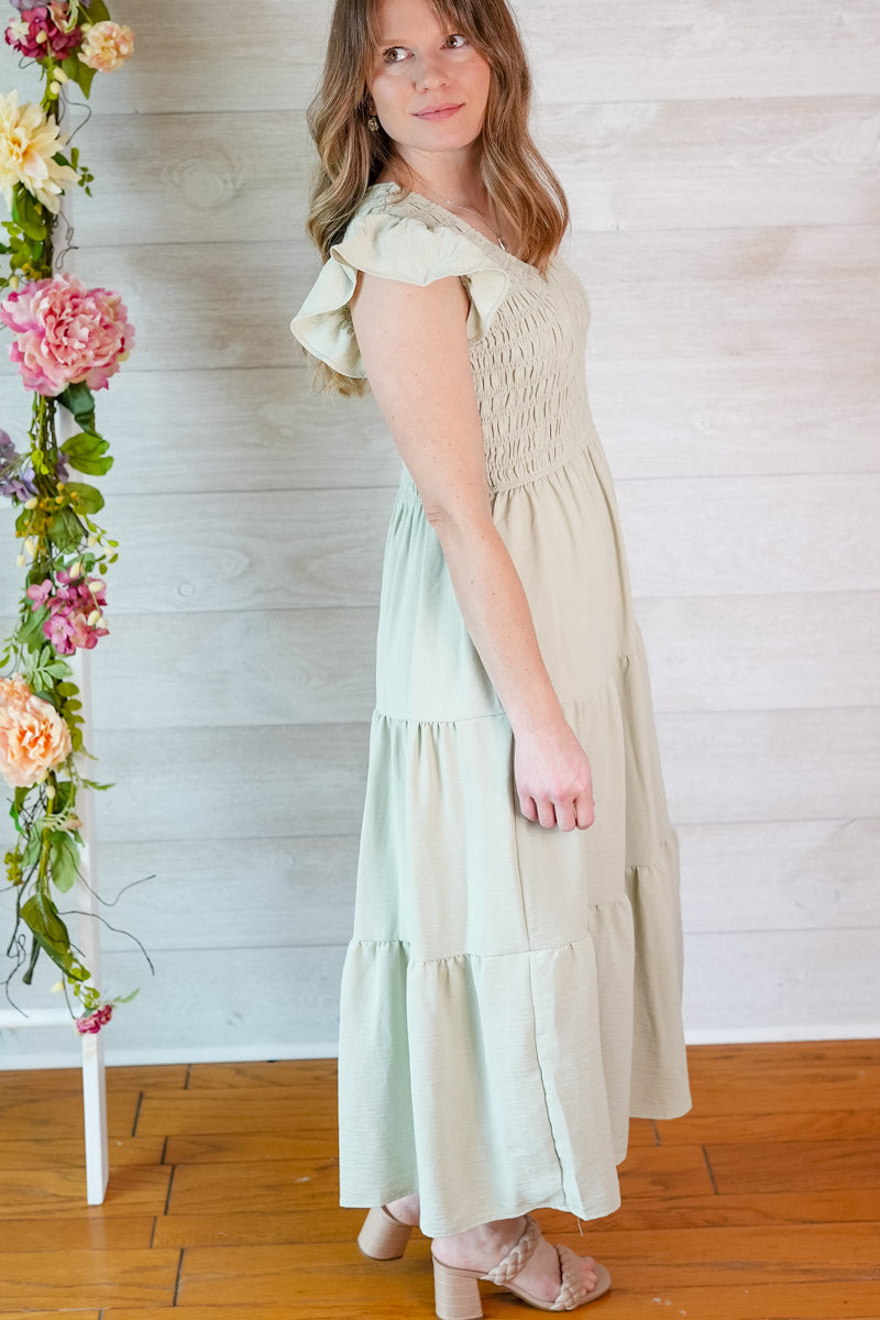 Woman wearing a light green dress standing next to a floral arrangement against a white wooden wall.