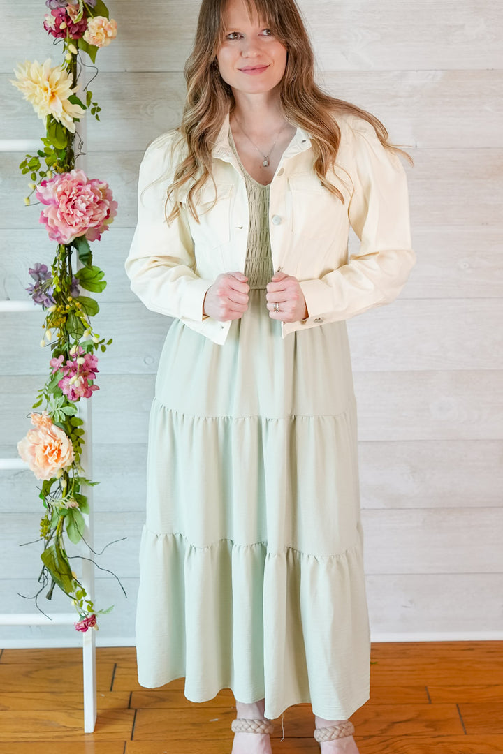 Woman in a light sage green colored dress and cropped jacket standing next to a floral arrangement against a wooden floor and white wall.