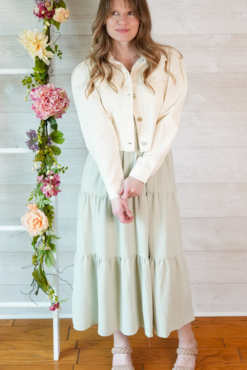 Woman in a light sage green colored dress and cropped puff sleeve jacket standing next to a floral arrangement indoors.