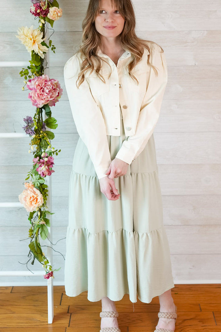 Woman in a light sage green colored dress and cropped puff sleeve jacket standing next to a floral arrangement indoors.