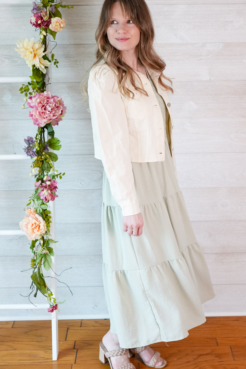 Woman in a light green colored dress and natural cropped puff sleeve jacket standing next to a floral arrangement against a white wooden panel background.