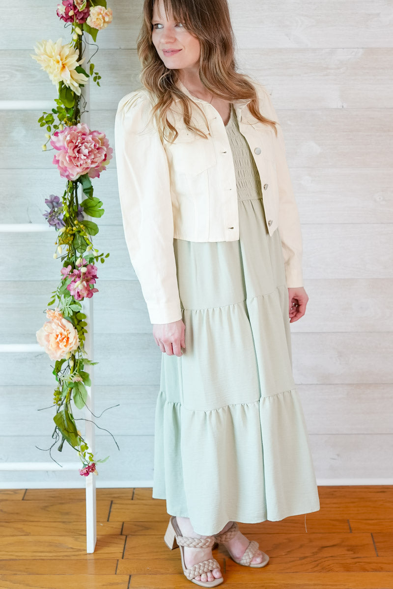 Woman in a light sage green colored dress and cropped puff sleeve jacket standing next to floral decorations indoors.
