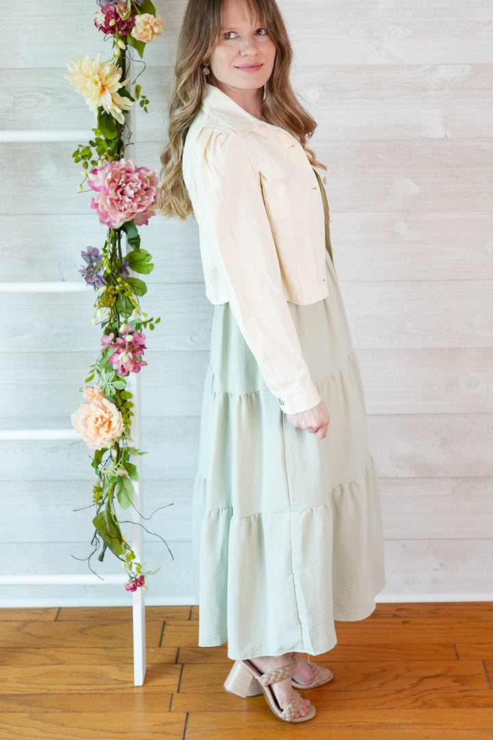 Woman in a light sage green colored dress  and cropped puff sleeve jacket standing next to a floral arrangement against a white wooden panel background