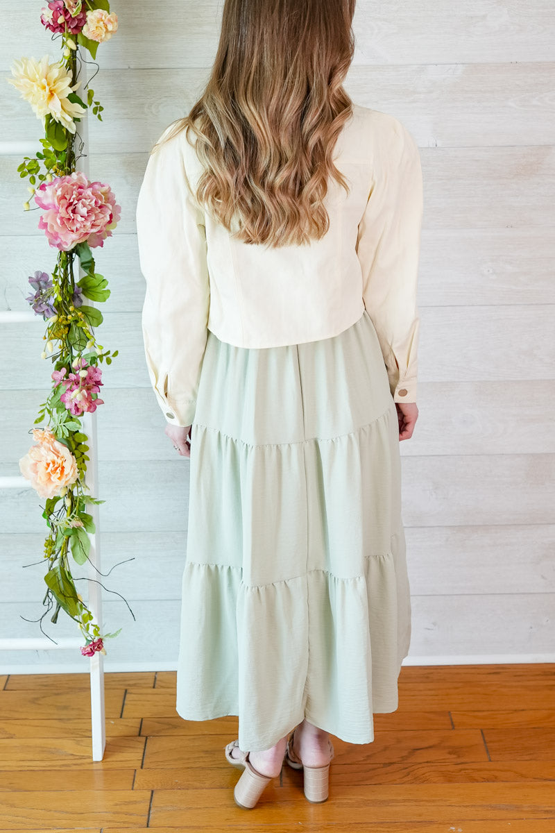 Woman wearing a light sage green colored dress and natural puff sleeve cropped jacket standing next to a floral arrangement against a wooden floor and white wall.