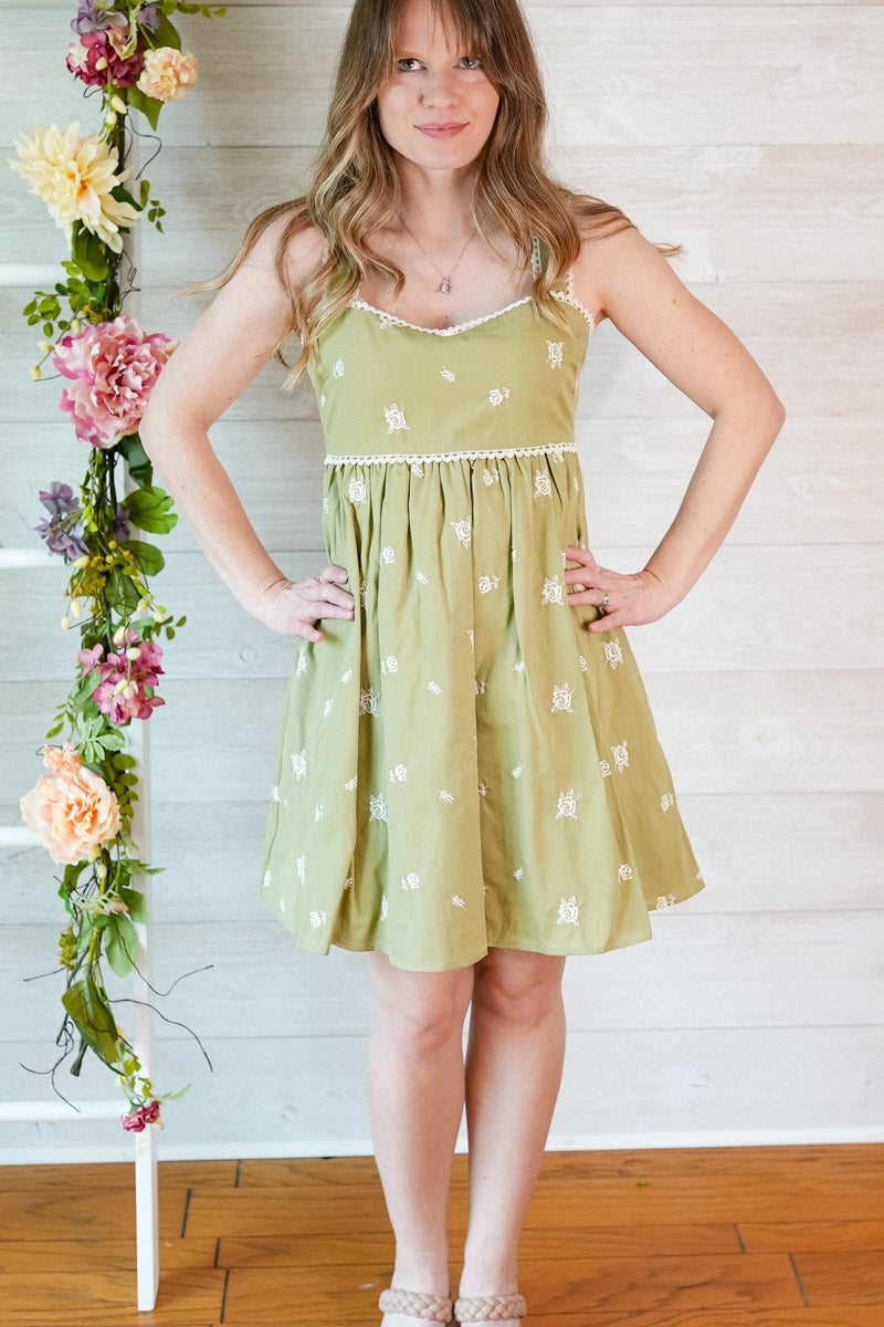 Young girl wearing a light green dress with floral patterns against a wooden floor and white wall background.