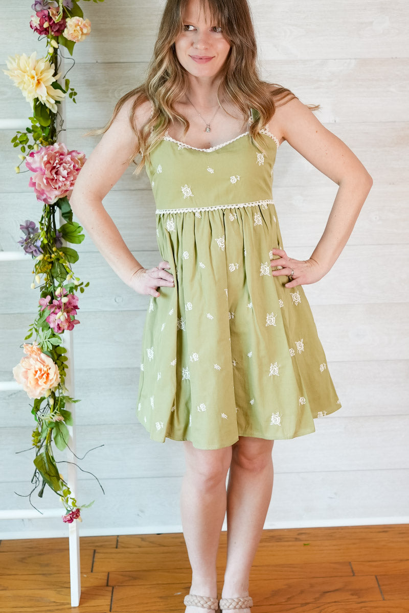 Woman wearing a green floral dress standing next to a floral arrangement against a white wooden panel background.