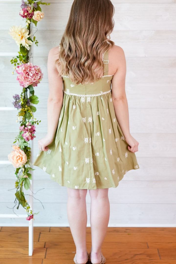 Woman wearing a green dress with white floral patterns, standing against a white wooden wall with floral decorations.