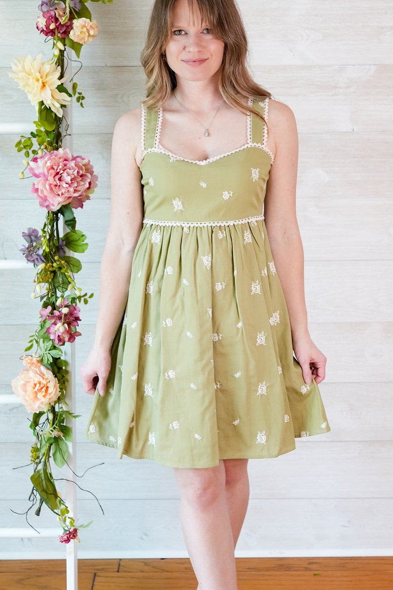 Woman wearing a green dress with white floral pattern standing next to floral decorations against a wooden wall.