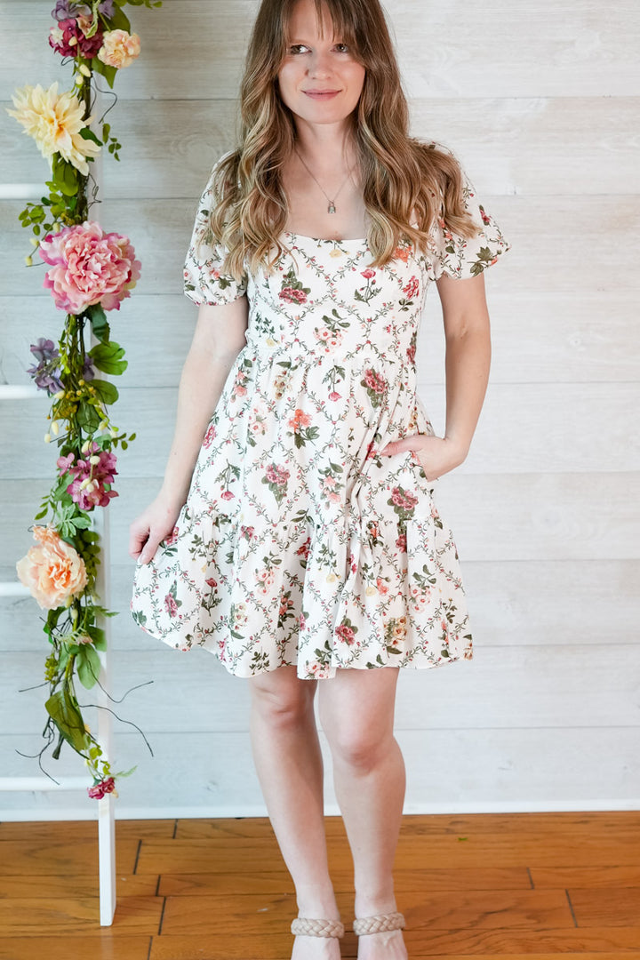 Woman wearing a short vintage floral dress standing next to a floral arrangement against a wooden floor and white wall.