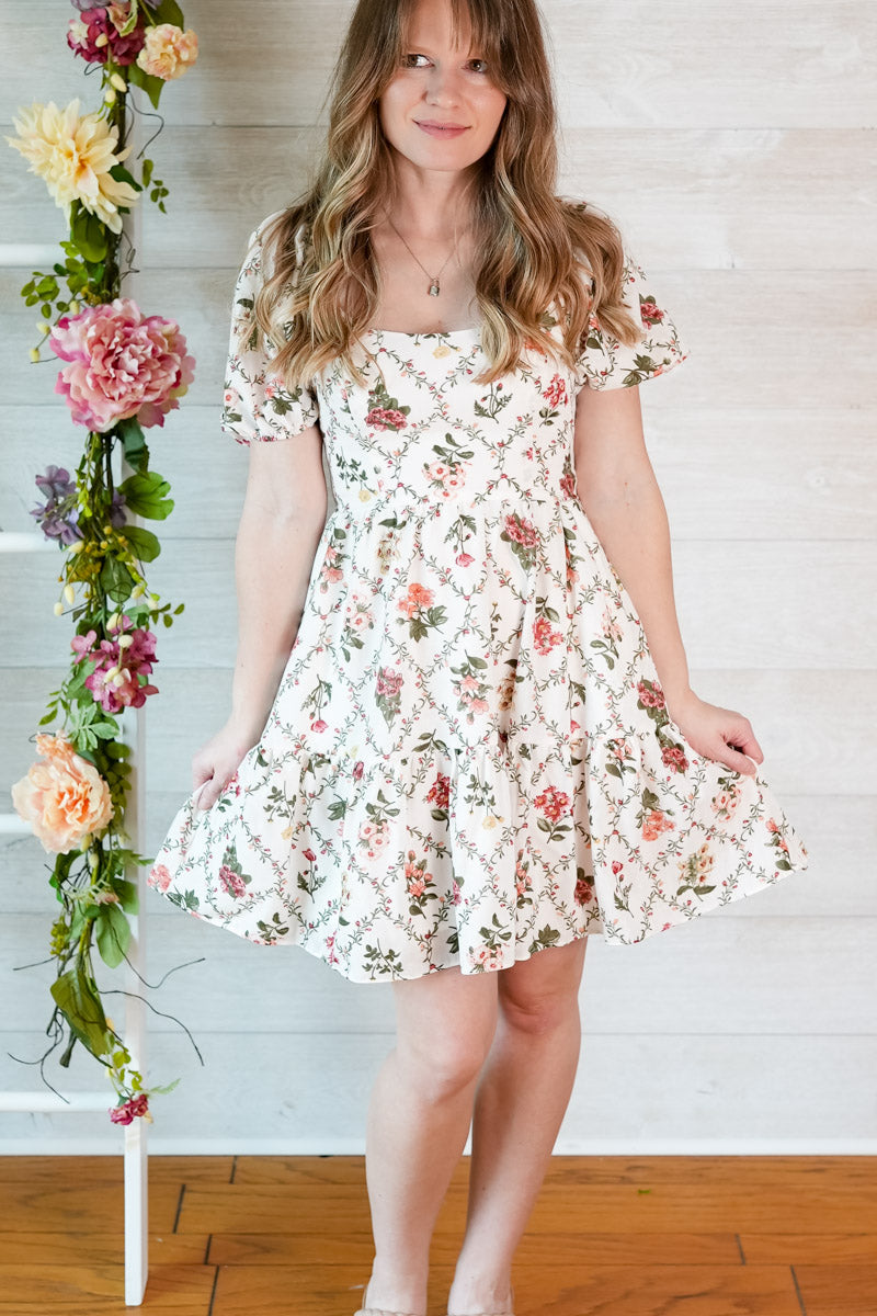 Woman wearing a floral dress standing next to a floral arrangement against a wooden floor and white wall.