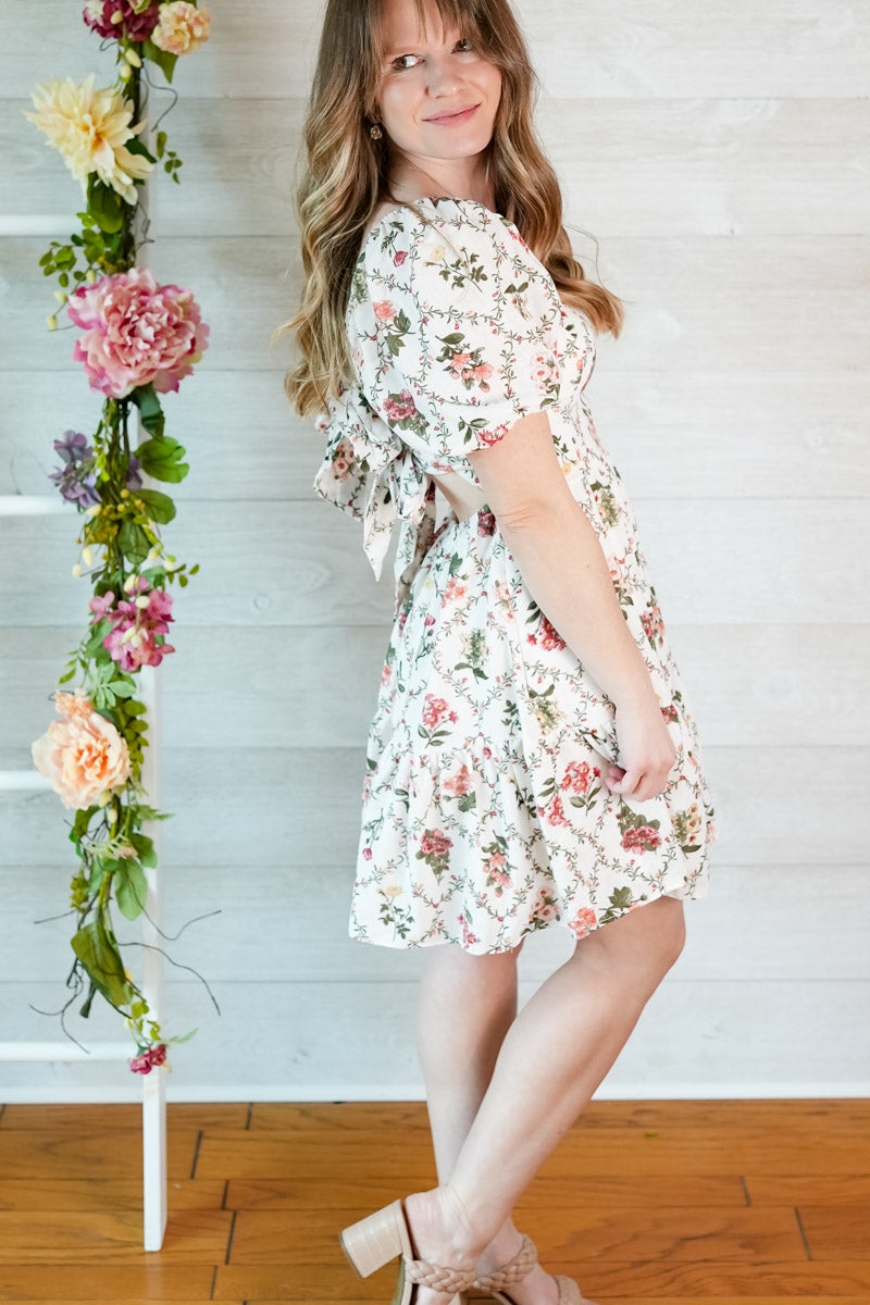 Young girl in a floral dress standing next to a decorative floral ladder against a white wooden panel wall.