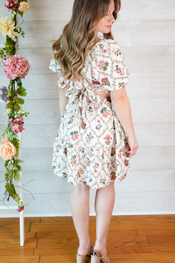 Woman wearing a floral dress standing next to a floral arrangement against a wooden floor and white wall.