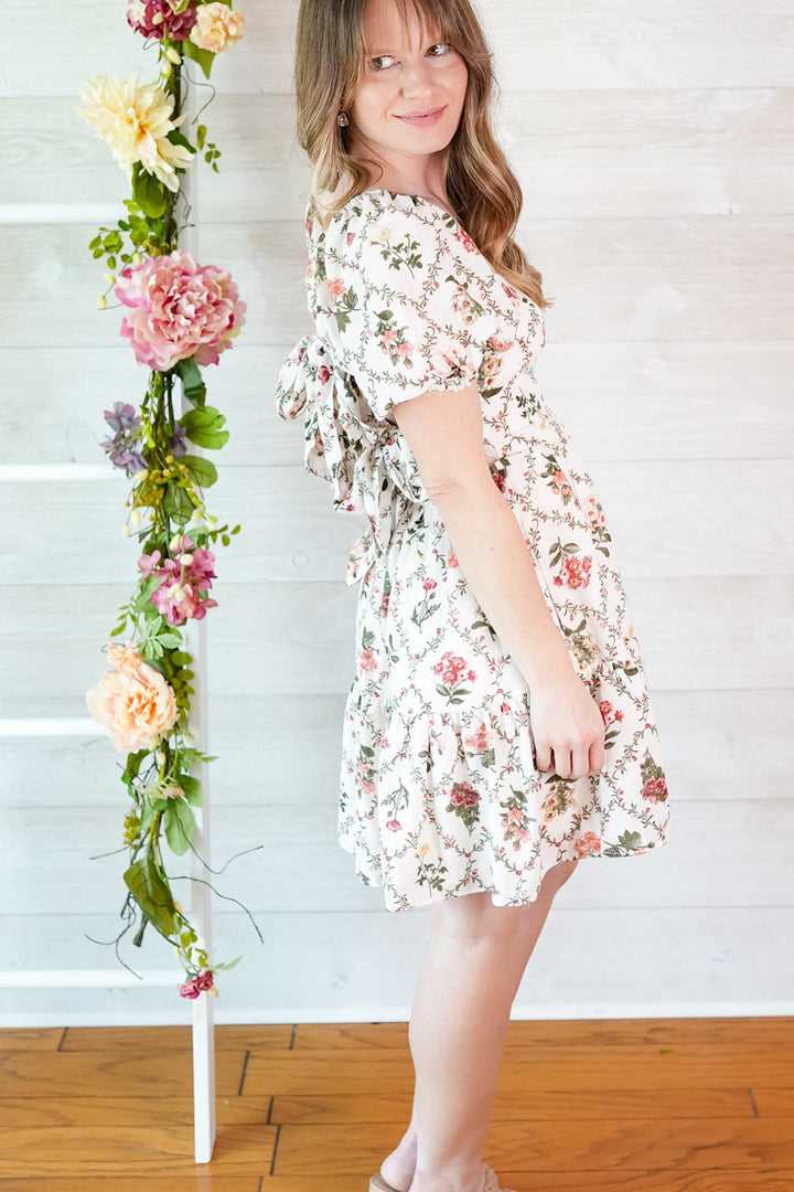 Woman in a floral dress standing next to a floral arrangement against a white wooden background