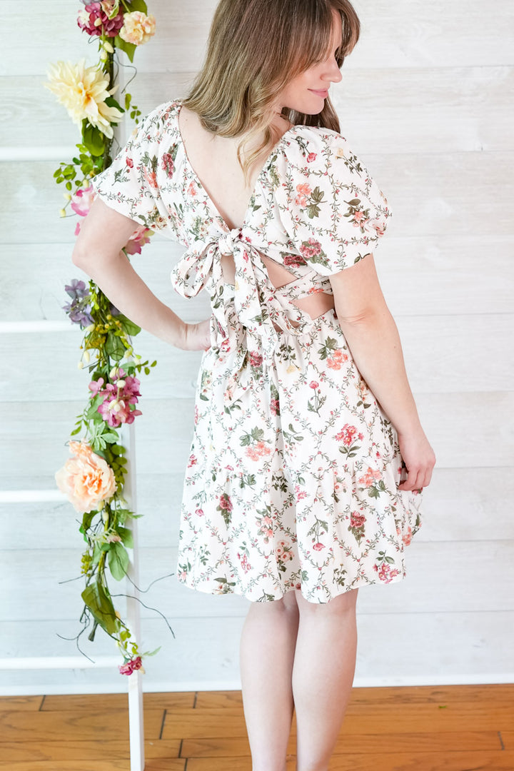 Woman wearing a floral dress standing next to a floral arrangement indoors.