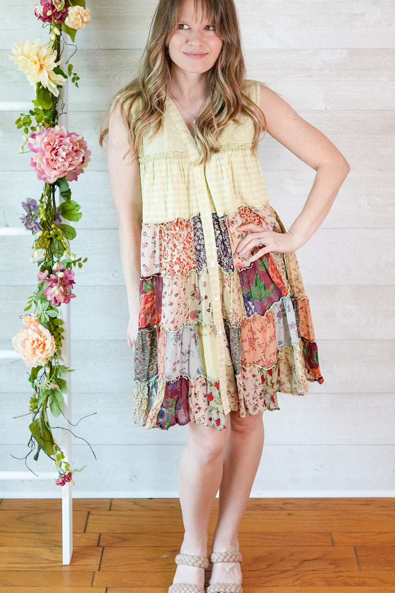 Woman wearing a colorful patchwork dress standing next to floral decorations indoors.