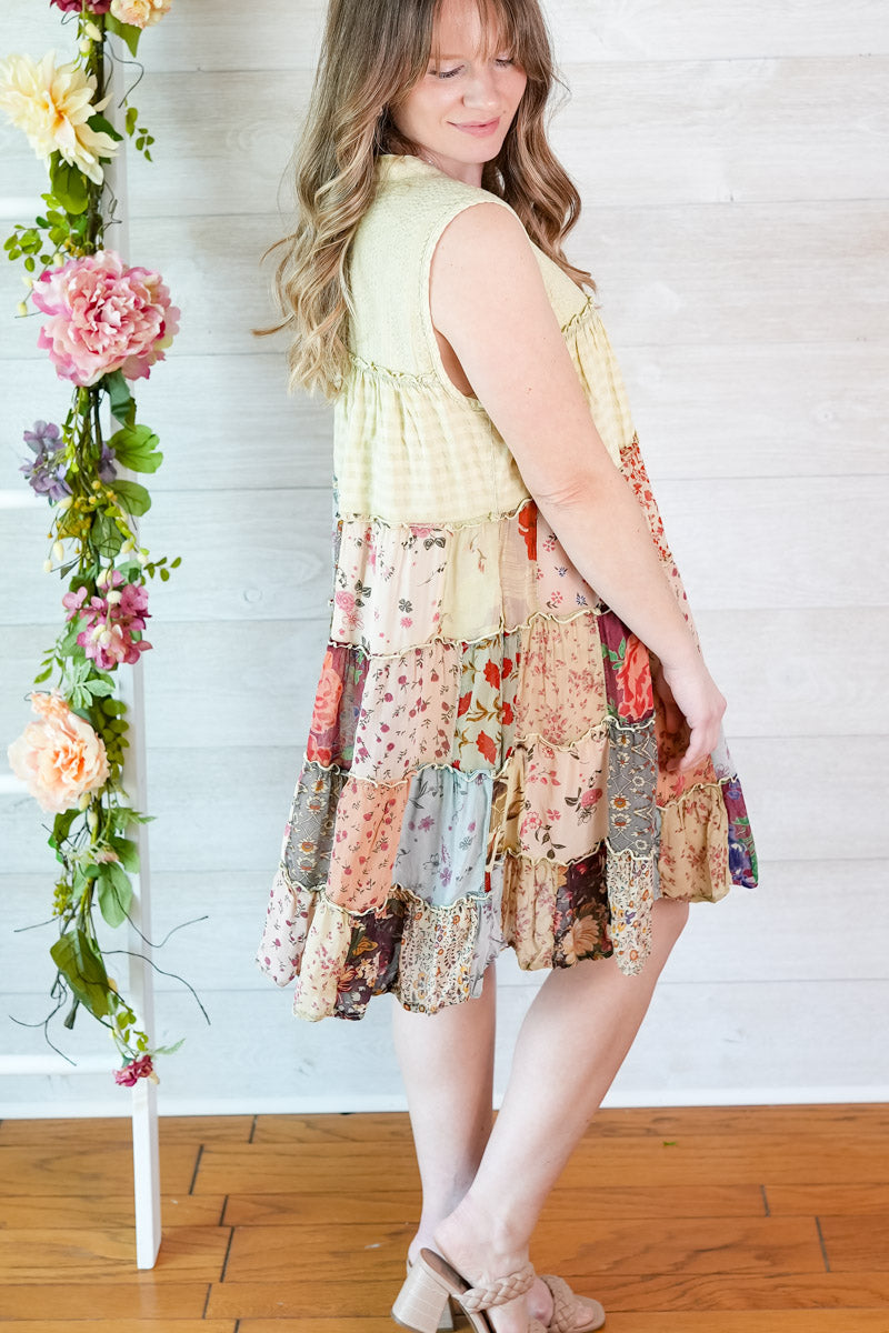 Woman wearing a colorful patchwork dress standing next to floral decorations against a white wall.
