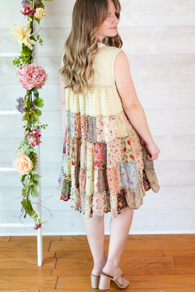 Woman wearing a patchwork dress standing next to a floral arrangement indoors.