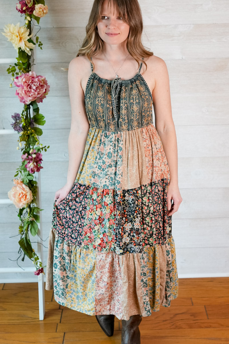 Woman wearing a patchwork dress with floral patterns, standing next to a floral arrangement indoors.