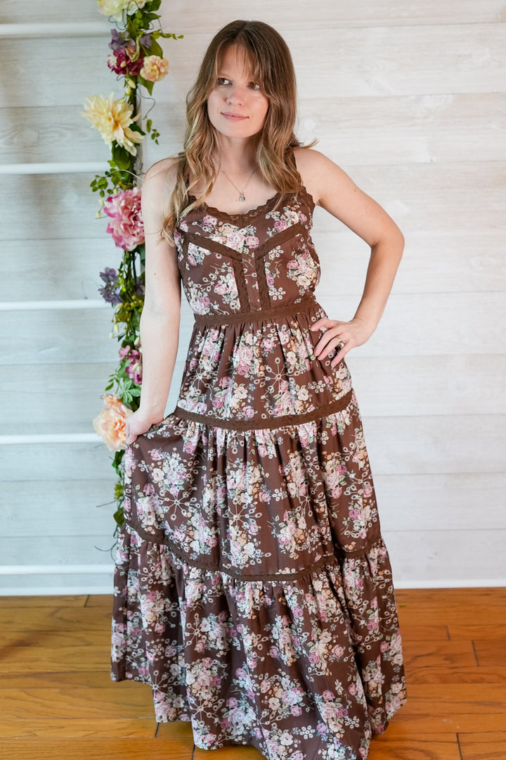 Woman wearing a brown floral western-inspired maxi dress standing in front of a white wall with decorative flowers.