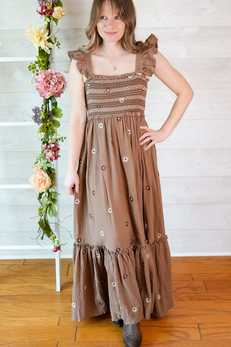 Woman wearing a brown floral dress standing next to a floral arrangement indoors.