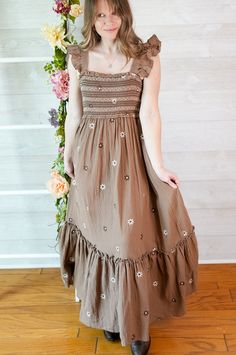 Woman wearing a brown dress with floral embellishments standing in front of a white wall with flowers.