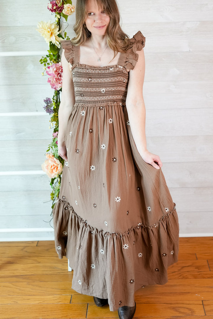 Woman wearing a brown dress with floral embellishments standing in front of a white wall with flowers.