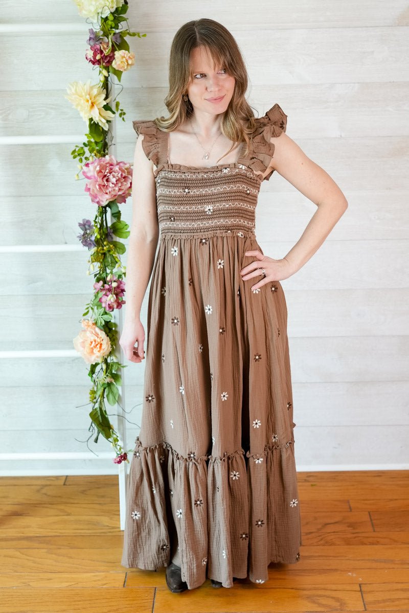 Woman wearing a western inspired brown floral embroidered maxi dress against a white wall with floral decorations.