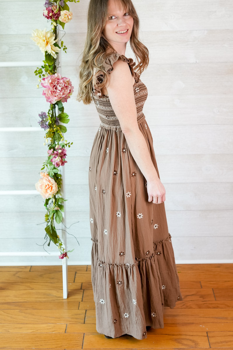 Woman wearing a brown dress with floral embellishments standing next to a floral arrangement indoors.