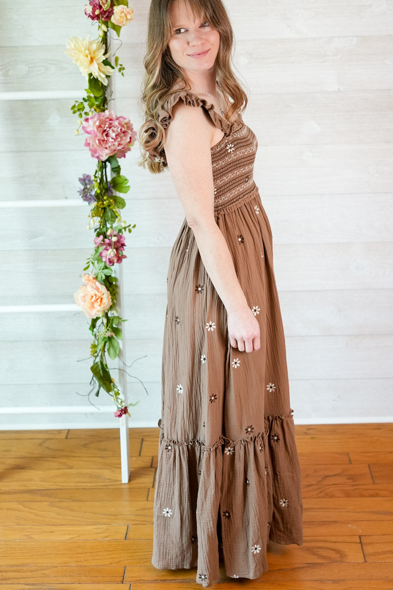 Woman in a brown flower embroidery maxi dress standing next to a floral arrangement indoors.