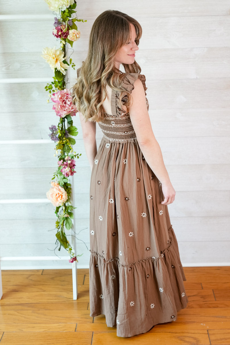 Woman wearing a western inspired floral embroidered brown maxi dress standing next to a floral arrangement indoors.