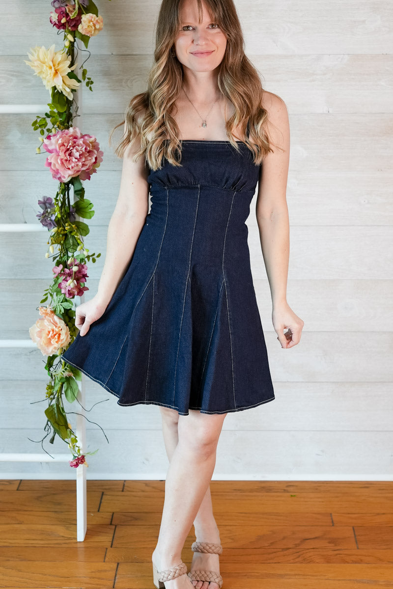 Woman wearing a dark denim blue fit and flare dress standing next to a floral arrangement indoors.