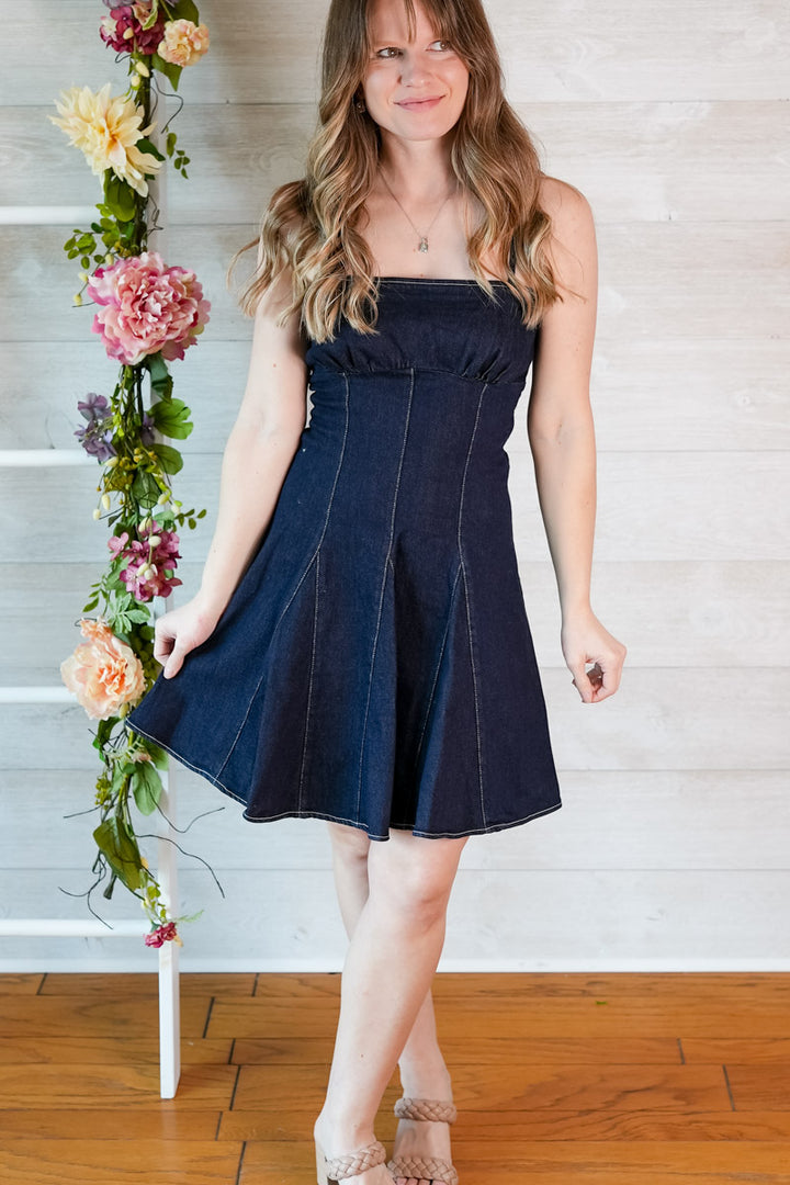 Woman wearing a dark blue denim fit and flare mini dress standing next to a floral arrangement indoors.