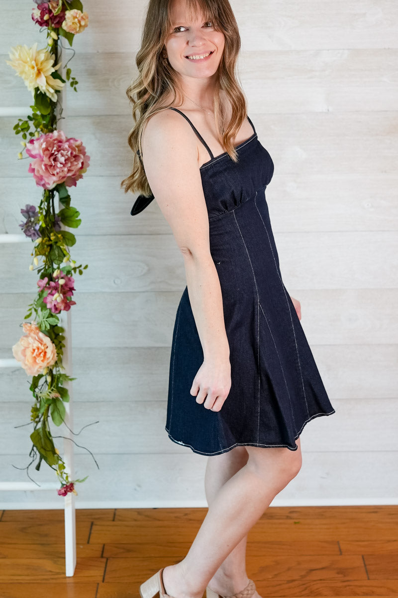 Woman in a dark blue denim mini dress standing next to a floral arrangement against a white wall.