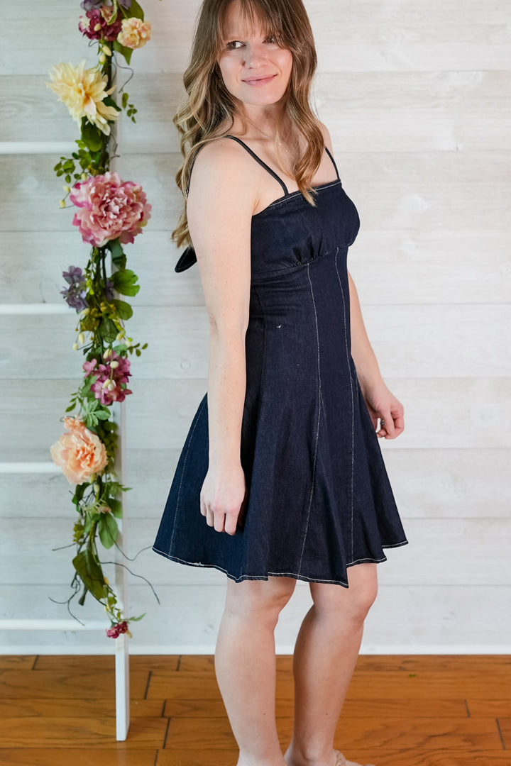 Woman wearing a dark blue denim mini dress standing next to a floral arrangement against a white wall.