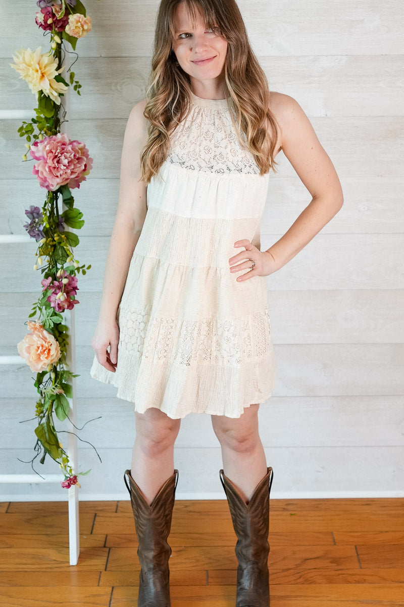 Woman wearing a neutral tiered halter dress with brown cowboy boots standing next to floral decorations.