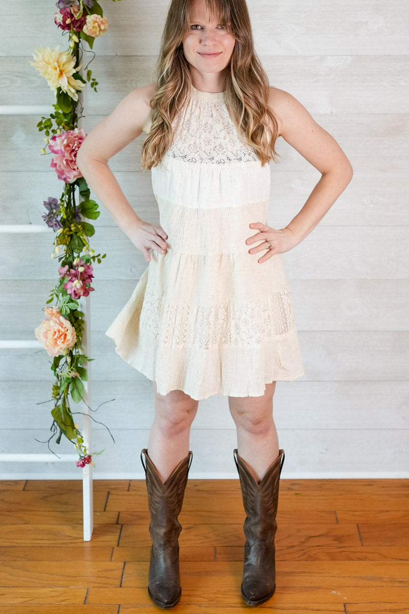 Woman wearing a white dress and brown cowboy boots standing in front of a floral arrangement.