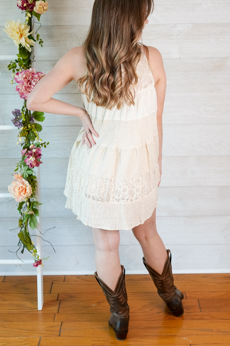 Woman in a white dress and brown boots standing next to a floral arrangement against a wooden floor and white wall.