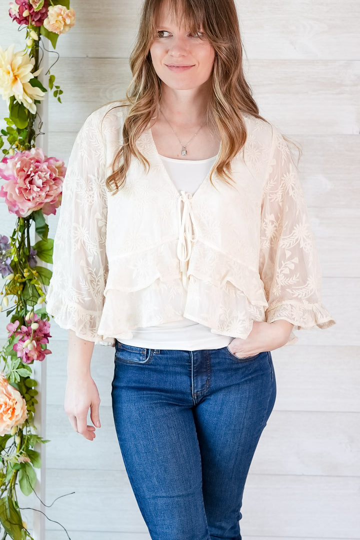 Woman wearing a light-colored blouse and blue jeans standing next to floral decorations.