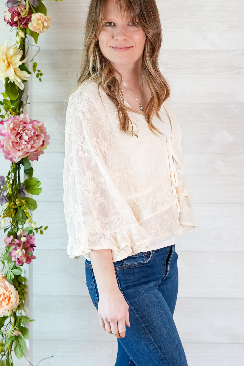 Woman wearing a light blouse and blue jeans standing next to floral decorations against a white wooden panel background