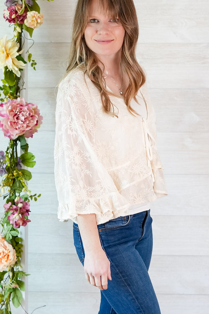 Woman wearing a light blouse and blue jeans standing next to floral decorations against a white wooden panel background