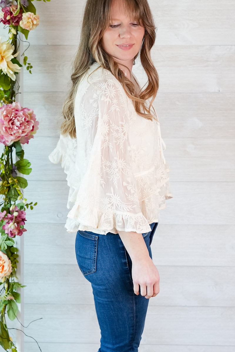 Woman wearing a white lace blouse and blue jeans standing next to floral decorations.