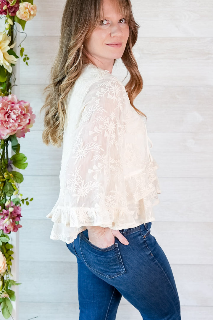 Woman wearing a white lace blouse and blue jeans standing next to floral decorations.