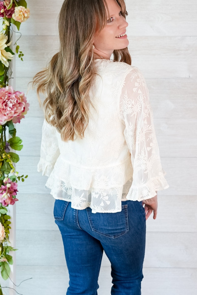 Woman wearing a white lace top and blue jeans standing next to floral decorations.